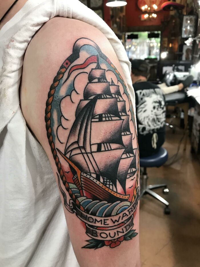 American traditional tattoo of a sailing ship on an arm, featuring bold colors and "Homeward Bound" banner.