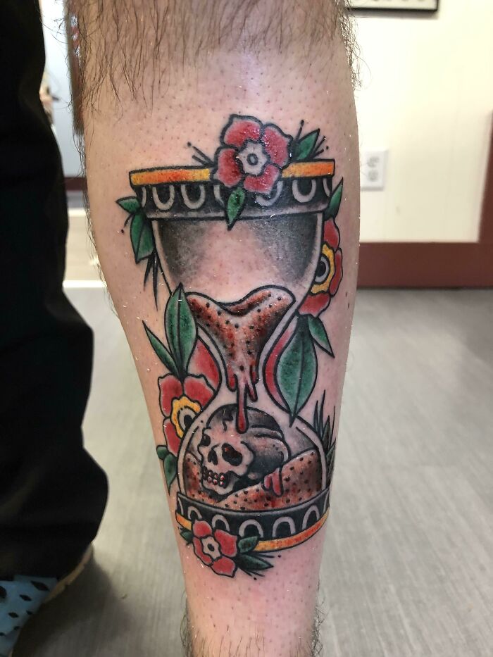 American traditional tattoo of an hourglass with flowers and a skull on a person's leg.