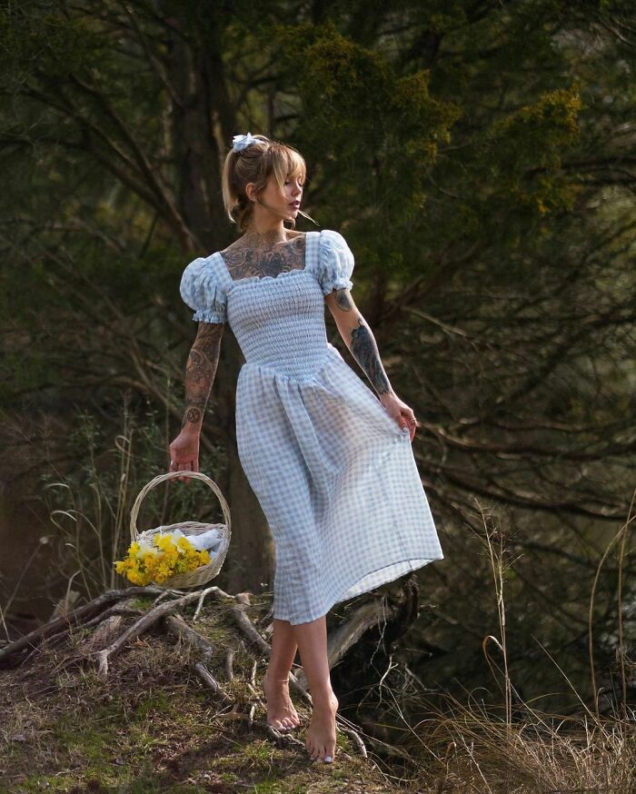 Woman with tattoos in a gingham dress, holding a basket of yellow flowers in a forest setting.