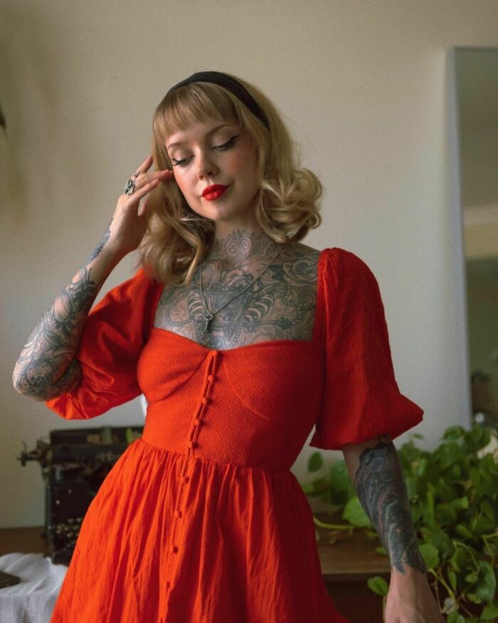A woman with regretful tattoos, wearing a red dress, stands indoors.