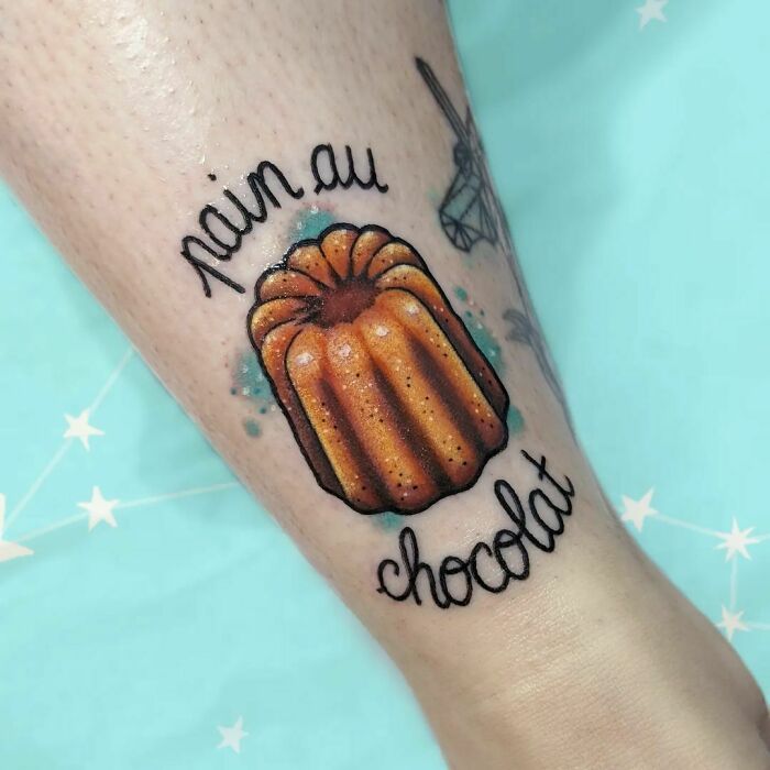 Watercolor cannelé tattoo with "pain au chocolat" text, showcasing food-inspired ink-spiration.