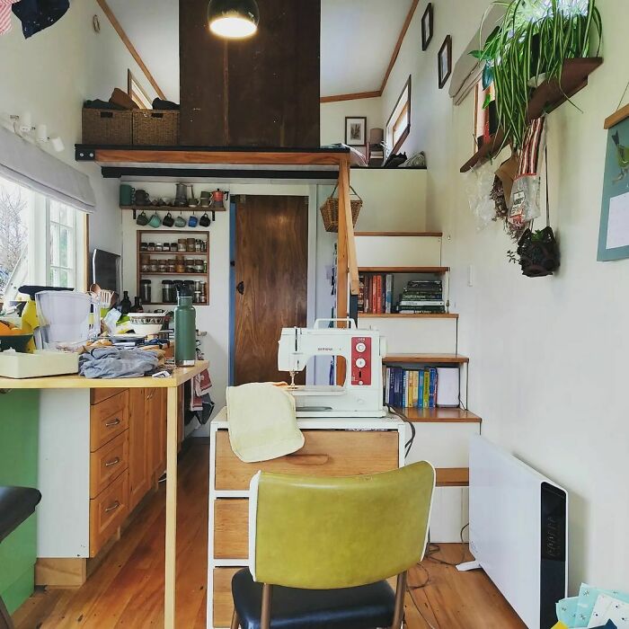 Cozy tiny house interior with white walls, brown kitchen cabinets, wooden stairs, plants, and a sewing machine workspace.