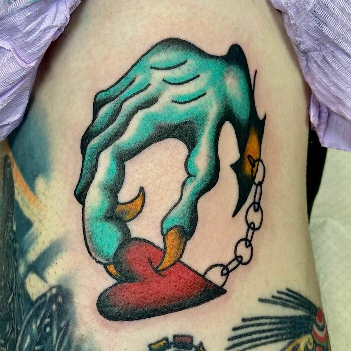 American traditional tattoo of a clawed hand holding a heart with a chain, showcasing bold colors and detail.