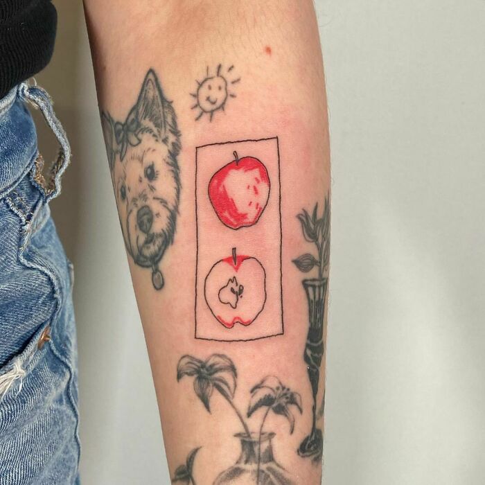 Watercolor apple tattoo on forearm with dog and plant designs, featuring bold red hues and natural inspiration.