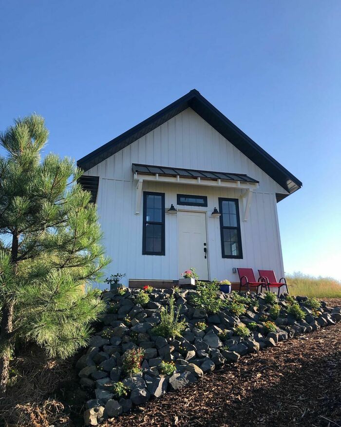 White small house with black trim surrounded by plants and rocks, showcasing a charming tiny house design outdoors.