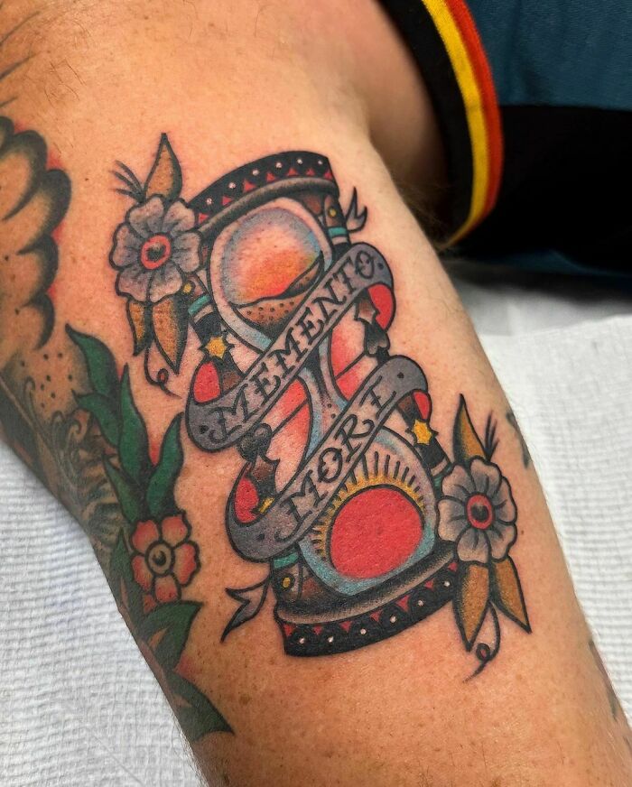 American traditional tattoo of an hourglass with "Memento Mori" banner, featuring flowers and vibrant colors.