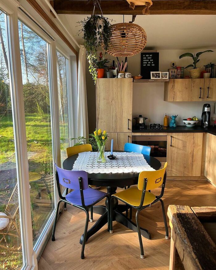 Cozy tiny house kitchen with wooden cabinets, colorful chairs, and a round dining table by large glass windows.