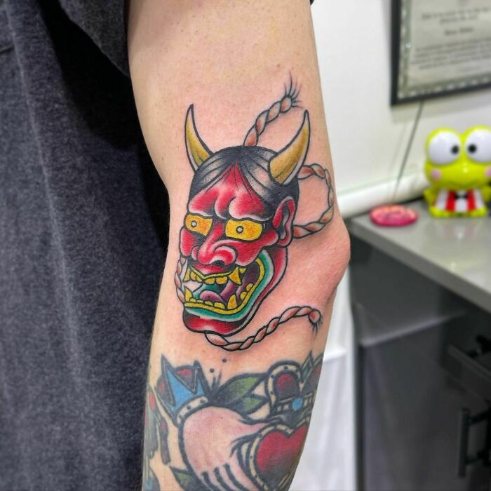 Colorful American traditional tattoo of a demon mask with yellow horns and braided hair on an arm.