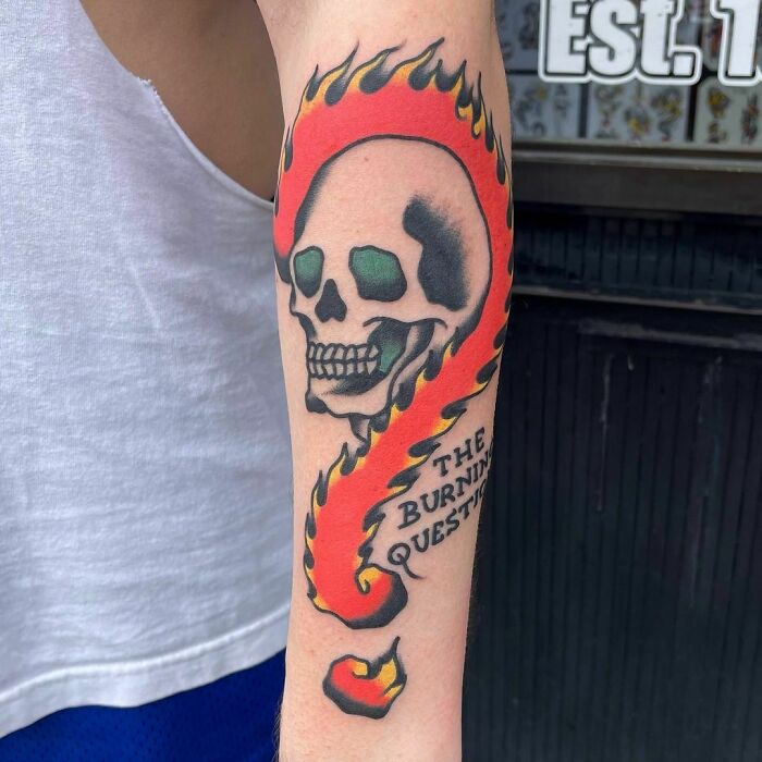 American traditional tattoo featuring a skull with flames and "The Burning Question" text on an arm.