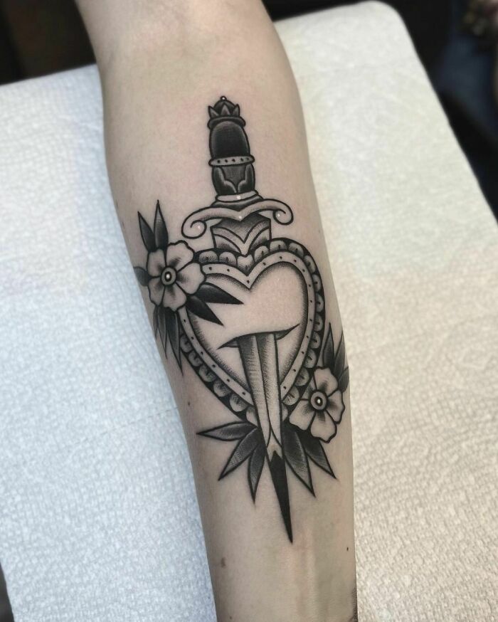 American traditional tattoo featuring a heart and dagger with floral elements on an arm.
