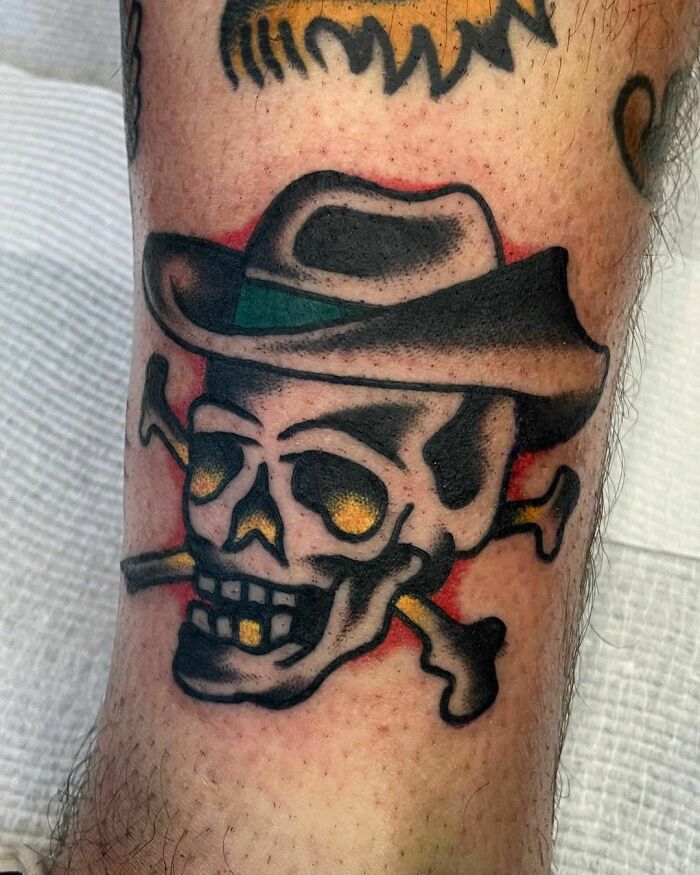 Skull and crossbones tattoo with hat, showcasing American traditional style on a leg.