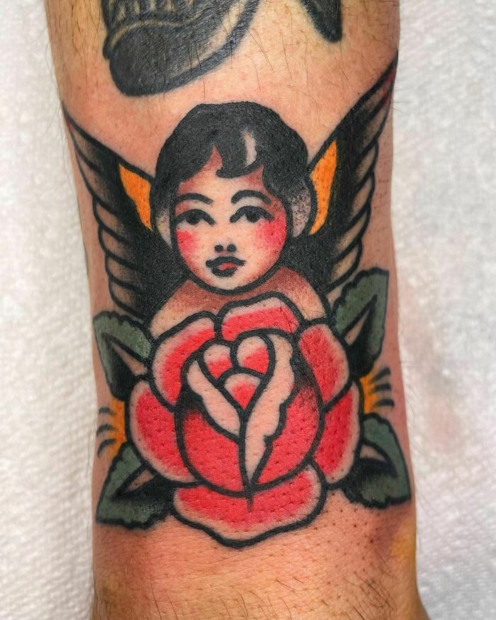 American traditional tattoo of a winged cherub above a red rose on skin.