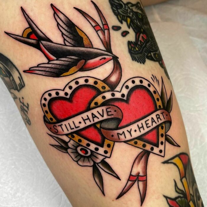 American traditional tattoo with two red hearts, a swallow, and a banner reading "Still have my heart" on a person's arm.