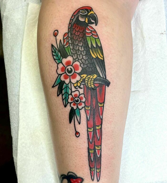 American traditional tattoo of a parrot with flowers in bold colors on forearm.