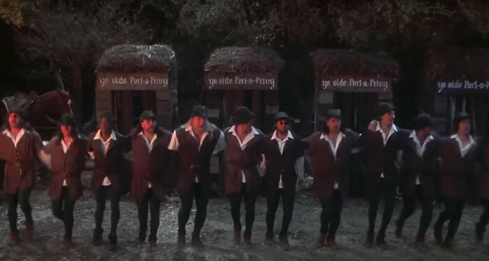 Many men in tights hold each other and dance in the forest