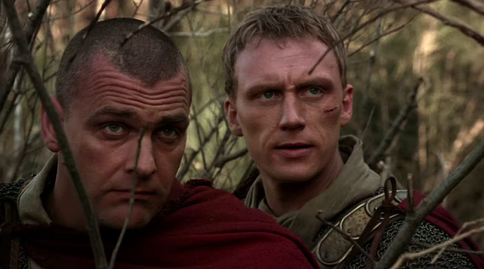 Two gladiators looking at something through bushes 