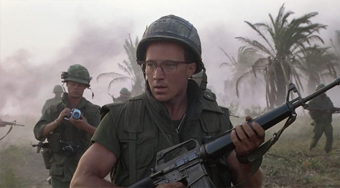Arliss Howard in a war zone holding a gun 