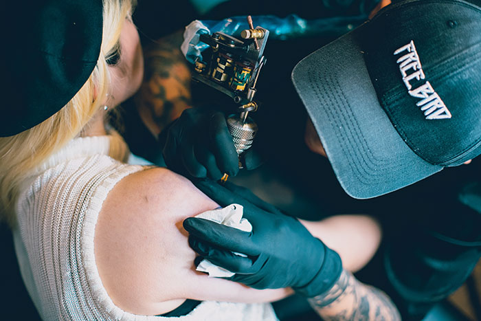 Tattoo artist working on a woman's shoulder tattoo, close-up view, black gloves and cap visible in the scene. Tattoo artist working on a woman's shoulder tattoo, close-up view, black gloves and cap visible in the scene.