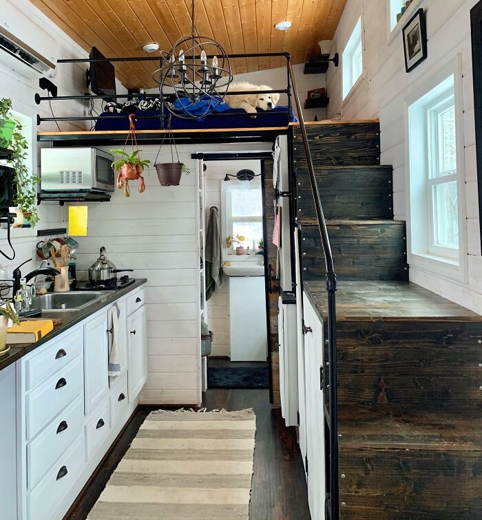 Cozy tiny house interior with a loft bed, wooden stairs, and a compact kitchen featuring white cabinets and a small sink.