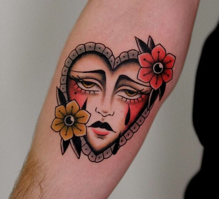 American traditional tattoo of a heart-shaped face with red and yellow flowers on an arm.