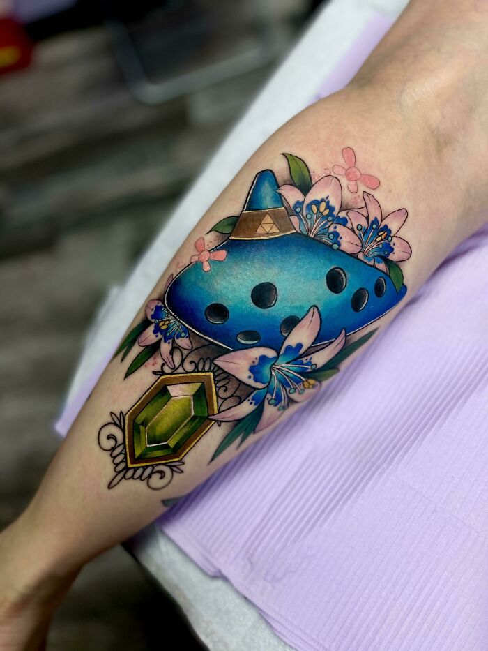 Colorful pop culture tattoo featuring an ocarina, flowers, and a green gem on forearm.