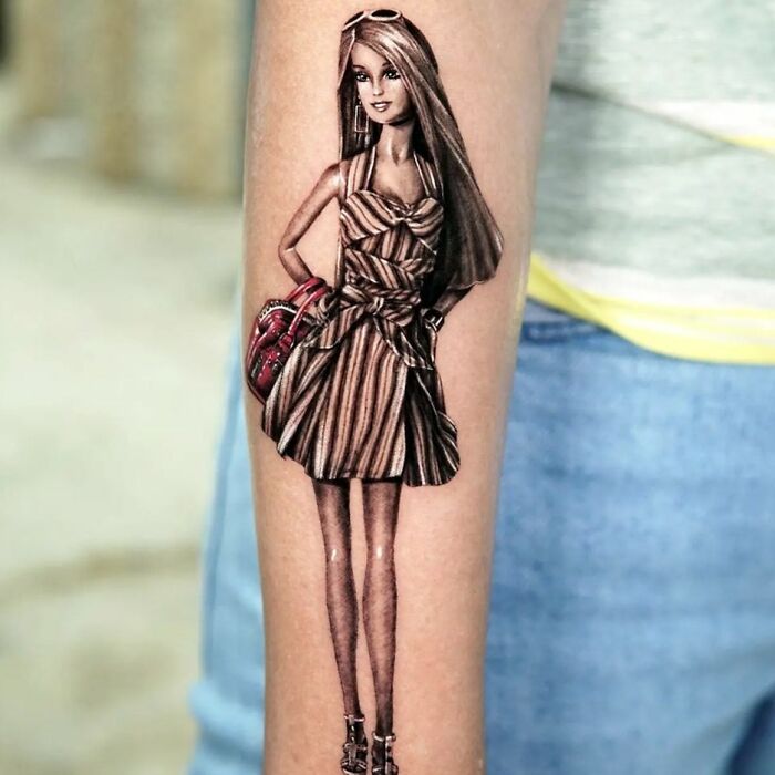 Pop culture tattoo of a fashionable doll wearing a striped dress, located on a person's arm.