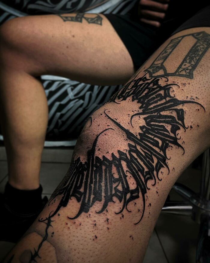 Ornate black ink knee tattoo design on a person's leg, showcasing bold, intricate lettering.