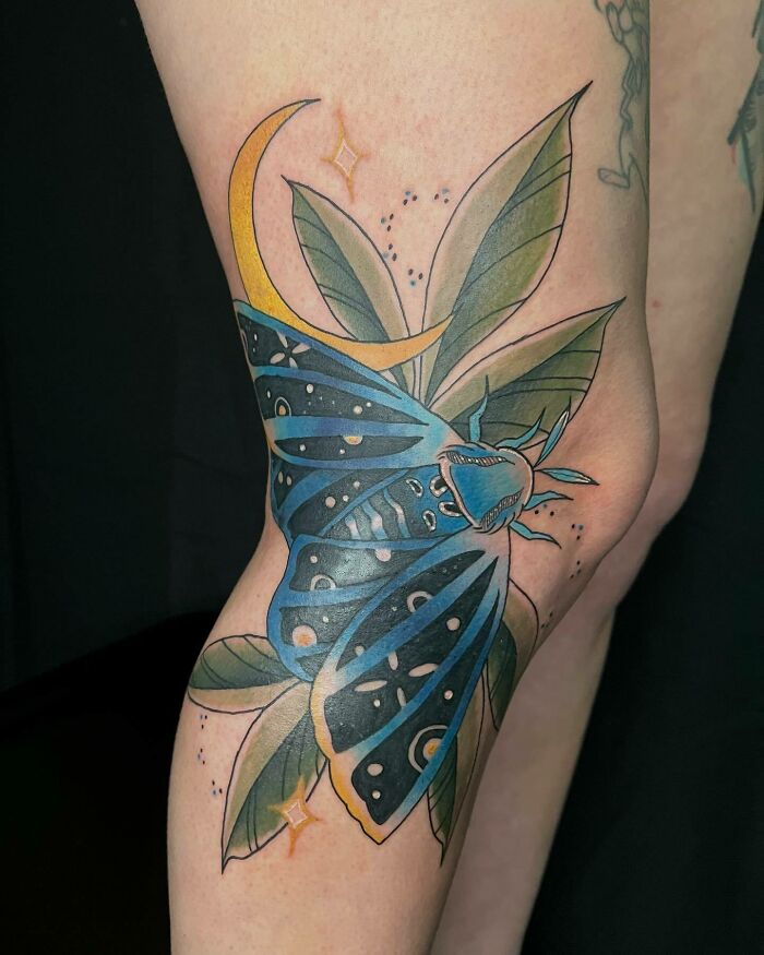 Colorful knee tattoo featuring a blue moth with green leaves and a yellow crescent moon.
