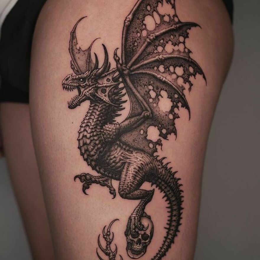 black ink tattoo of a flying dragon with a skull on its foot