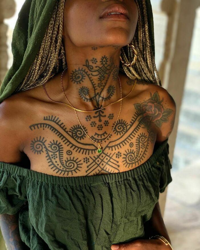 Woman with intricate tribal tattoos on chest and neck, wearing a green off-shoulder top and gold necklaces.