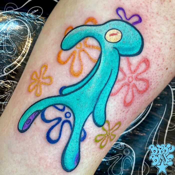 Pop culture tattoo featuring a blue abstract character with colorful flower designs on skin.