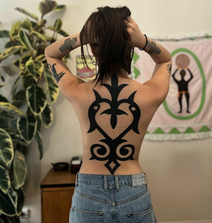 Person with a large tribal tattoo on their back, standing in front of wall art and plants.