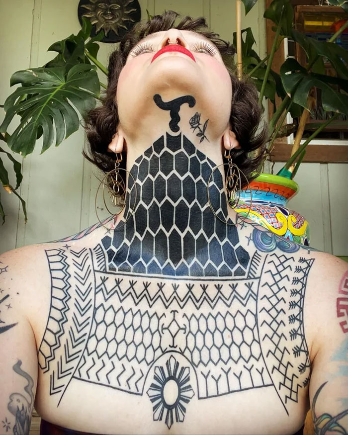 Person showcasing intricate tribal tattoo designs on neck and chest against a backdrop of indoor plants.