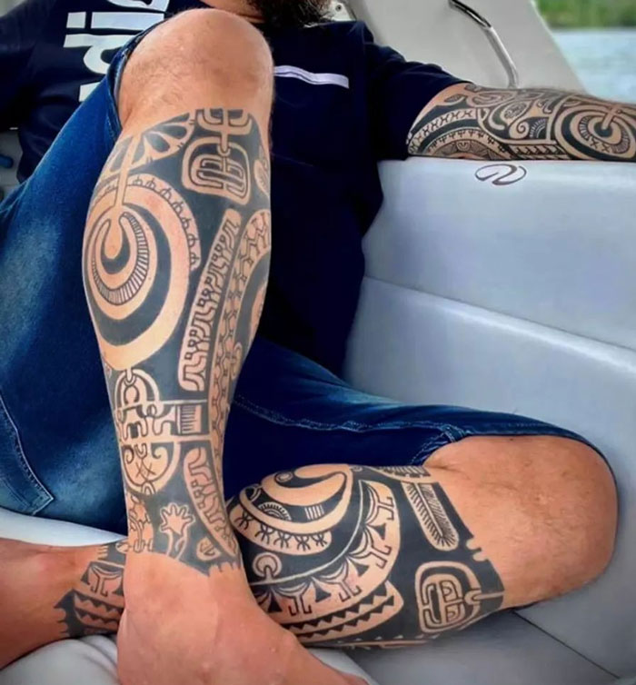 Man with intricate tribal tattoos on arm and legs, wearing denim shorts, seated casually on a boat.