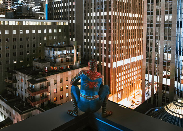 Person dressed as Spider-Man crouching on a rooftop overlooking city buildings at night in a college courses setting.