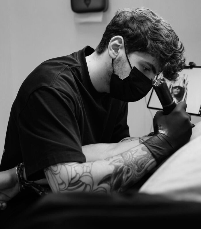 Tattoo artist creating geometric design; black mask, gloves, and detailed arm tattoos visible. Tattoo artist creating geometric design; black mask, gloves, and detailed arm tattoos visible.