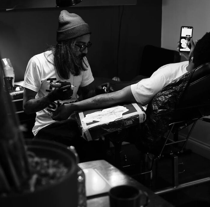 Tattoo artist working on a geometric tattoo, wearing a beanie and glasses, in a dimly lit studio. Tattoo artist working on a geometric tattoo, wearing a beanie and glasses, in a dimly lit studio.
