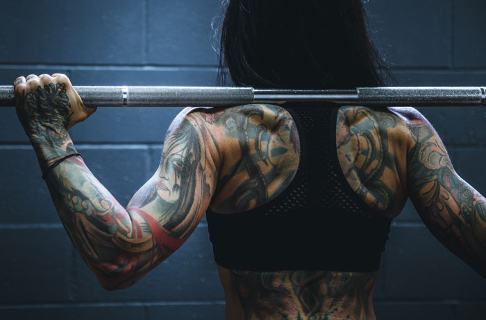 Person with colorful tattoos lifting a barbell, showcasing detailed body art against a dark background.
