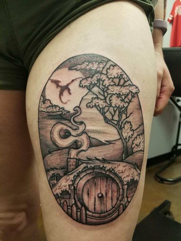 Lord of the Rings tattoo featuring a Hobbiton scene with a dragon, detailed on a person's thigh.