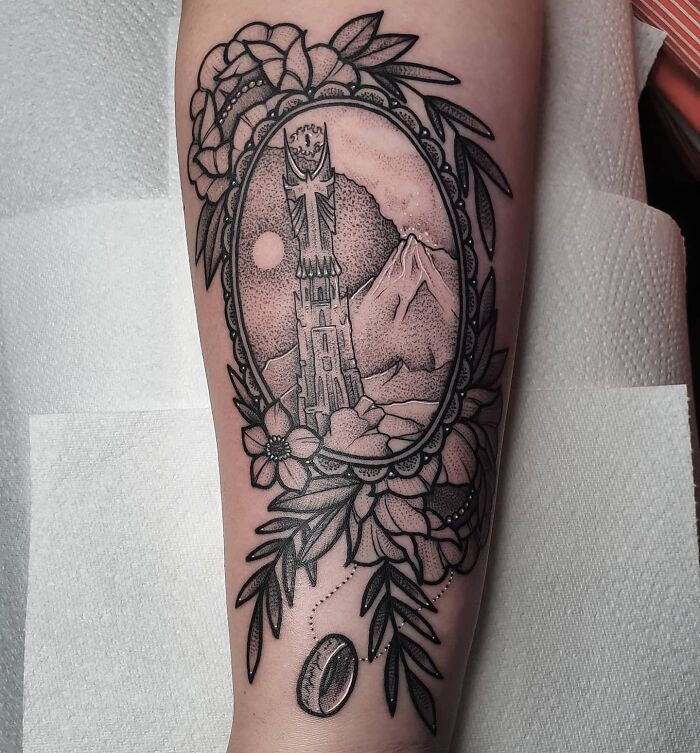Lord of the Rings tattoo featuring a tower and mountain, framed by flowers and leaves on an arm.