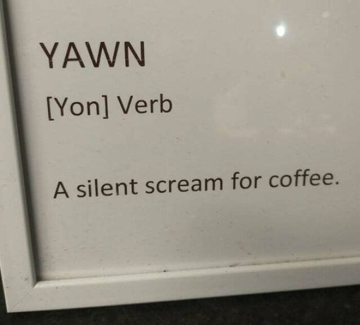 A coffee meme featuring a framed sign that humorously defines the word "Yawn" as "[Yon] Verb: A silent scream for coffee." The meme playfully suggests that yawning is a way of expressing the need for coffee, likening it to a cry for help in the form of caffeine.