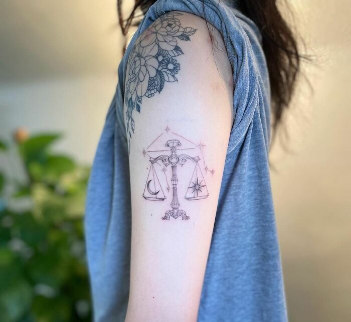 Zodiac tattoo of Libra scales with moon and star details on upper arm against a blurred background.
