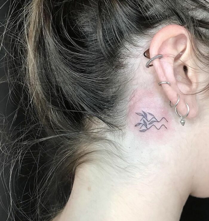 Minimalist zodiac tattoo behind ear with simple lines and wave elements.