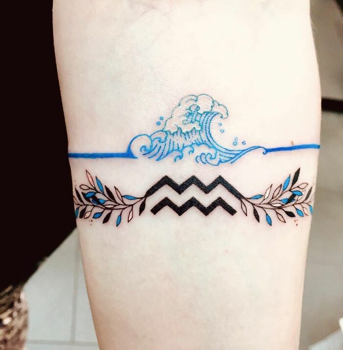 Aquarius zodiac tattoo with waves and decorative branches on forearm.