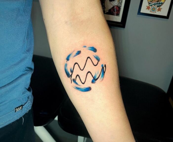 Aquarius zodiac tattoo with blue accents on forearm, showcasing bold, artistic design.