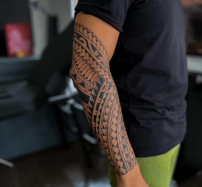 Arm with intricate tribal tattoo design in black and grey, showcased in a casual indoor setting.