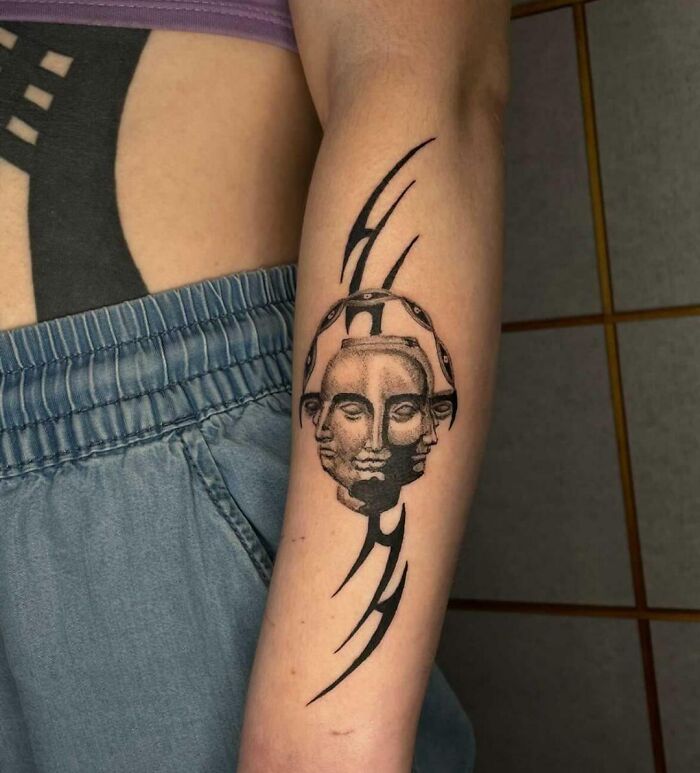 Arm with tribal tattoo featuring a double-faced figure and curved lines, worn with blue shorts.