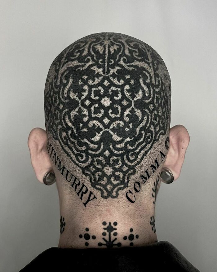 Ornate gothic tattoo covering the back of a shaved head with intricate patterns and black lettering.