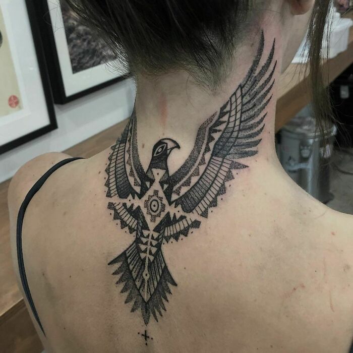 Tribal tattoo of a stylized bird on the back of a woman's neck in a well-lit room.