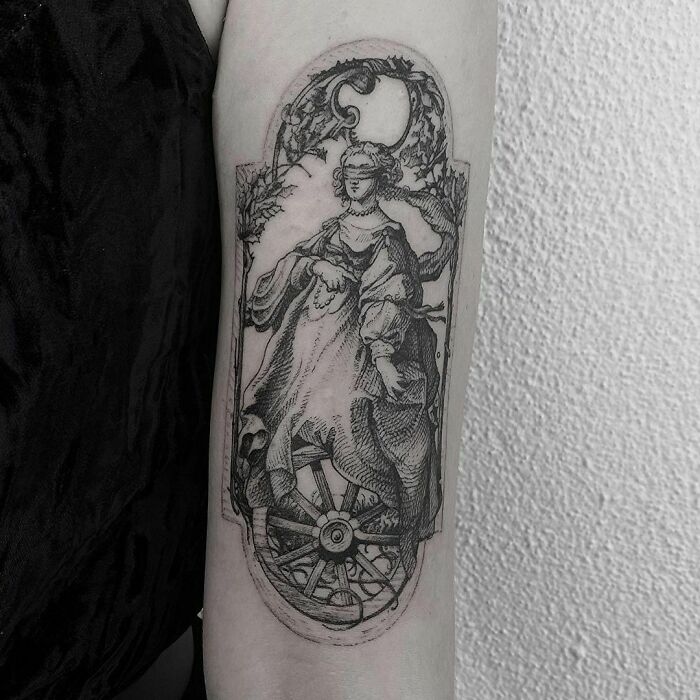 Black and white gothic tattoo depicting a classical figure with a skull and wheel detail on forearm.